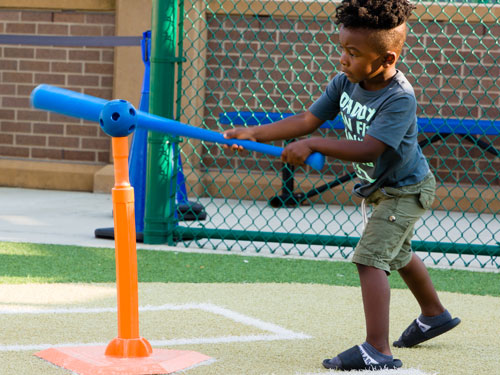 Child holding a plastic baseball bat up to a whiffle-style ball that's sitting on an orange baseball tee.